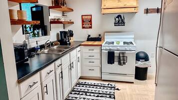 Une cuisine de taille réduite avec des meubles blancs, une cuisinière, un évier et un réfrigérateur. On trouve des étagères en bois au-dessus du plan de travail et une poubelle noire sur le côté.
