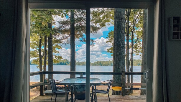 Une terrasse en bois avec une table et des chaises, donnant sur un lac bordé d’arbres et surplombant un ciel nuageux.