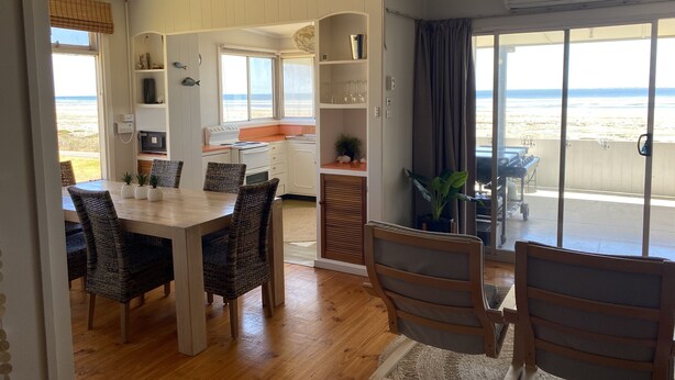 A dining area with a wooden table and wicker chairs, a kitchen with white cabinets, and a view of the beach through large windows. - Classic Shack -  Absolute Beachfront.  Winter off peak special! (Point Turton)