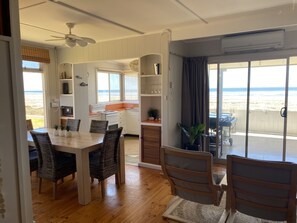 A dining area with a wooden table and wicker chairs, a kitchen with white cabinets, and a view of the beach through large windows. - Classic Shack - Absolute Beachfront. Winter off peak special! (Point Turton)