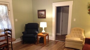 A living room with a wooden floor, a black armchair, a beige armchair, a wooden side table with a lamp, and a framed picture on the wall.