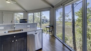 Ett modernt kök med en stor köksö, rostfria vitvaror och utsikt över ett hus och träd genom stora fönster.