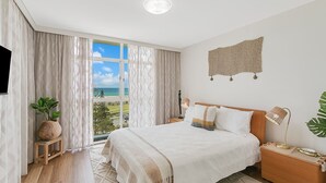 Un dormitorio con una cama grande, vista al océano, una mesita de noche de madera, una lámpara y un adorno en la pared.