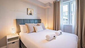 A neatly made bed with white linens and two rolled towels, a headboard, a bedside table with a lamp, and a window with curtains. - Stylish Harbour View Apartment in a Private Building (Galway City)
