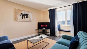 A modern living room with a sofa, a coffee table, a TV displaying 'NETFLIX', and a city view through large windows. - Stylish Harbour View Apartment in a Private Building (Galway City)