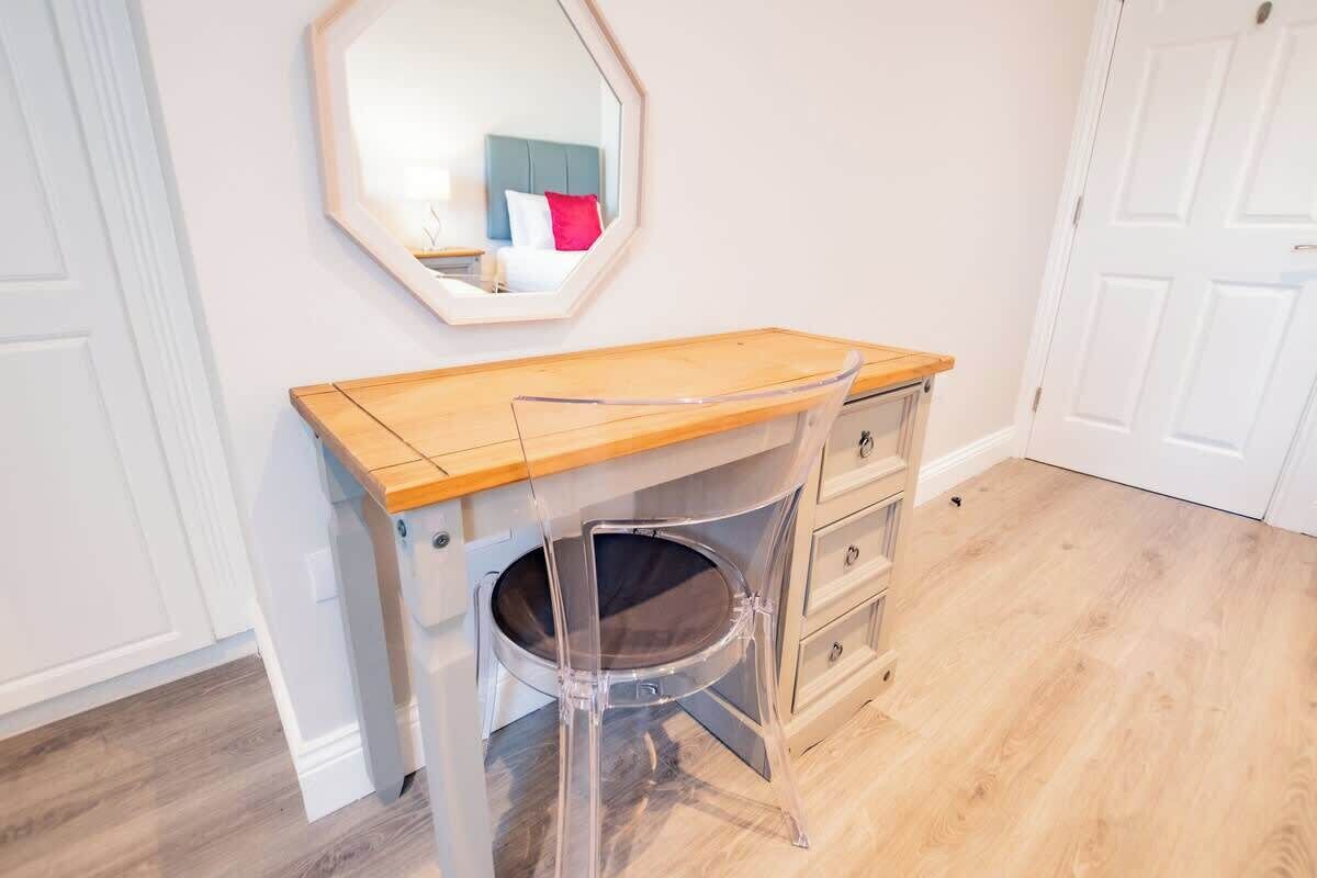 A room with a wooden desk, a chair with a transparent backrest, a mirror on the wall, and a white door.