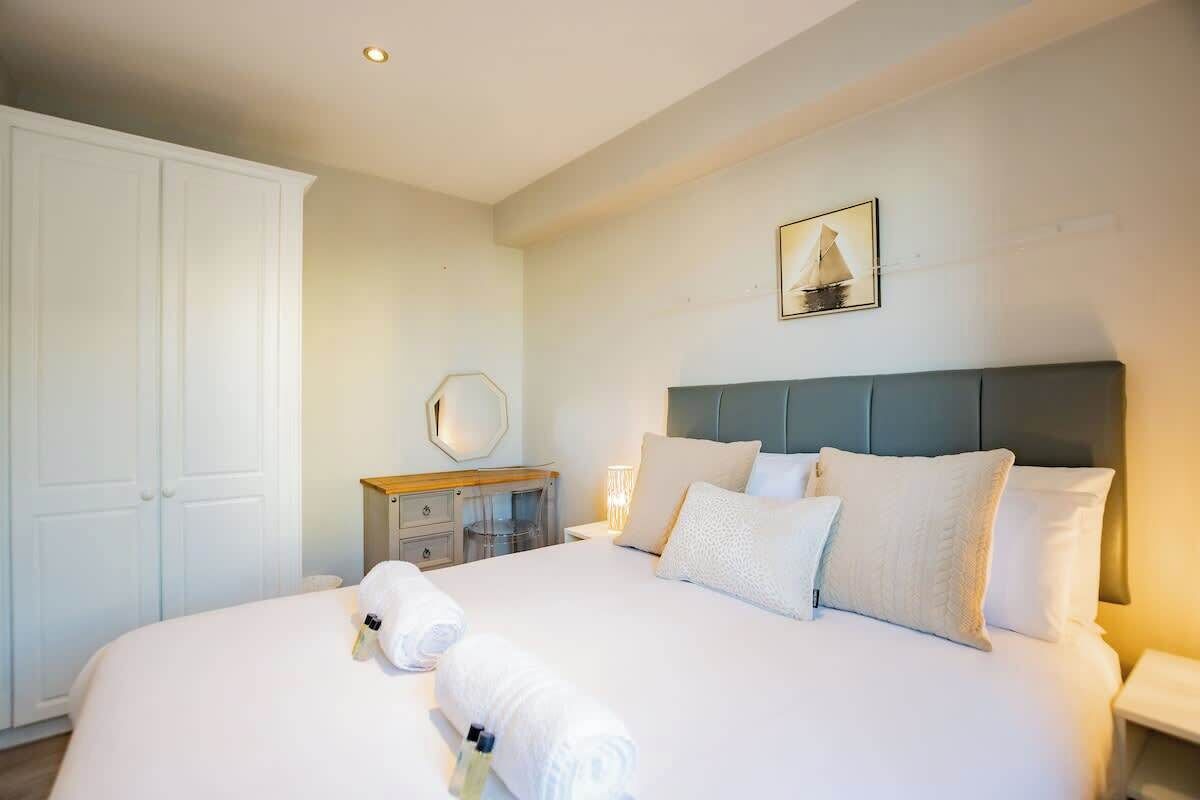 Una cama bien hecha con sábanas y almohadones blancos, una mesita de noche con lámpara, un armario y un cuadro en la pared.