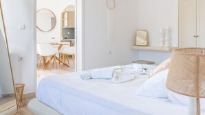 A bedroom with a bed, bedside table, lamp, and a mirror. There is a door leading to a bathroom and a desk with chairs. - Premium Pool Villa for 4 guests (Patmos)