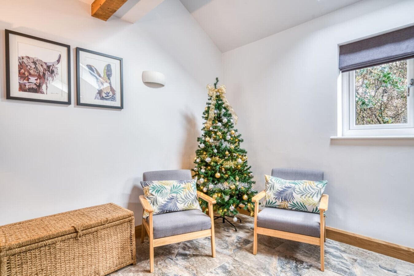 Una habitación de esquina con un árbol de Navidad, dos sillones, una cesta de mimbre y cuadros en la pared.