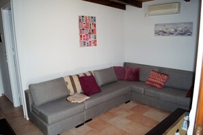A grey sofa with patterned cushions, a tiled floor, and a wall with a framed picture and a light fixture. - Holiday home in Historical village of Kefalonia (Troianata)