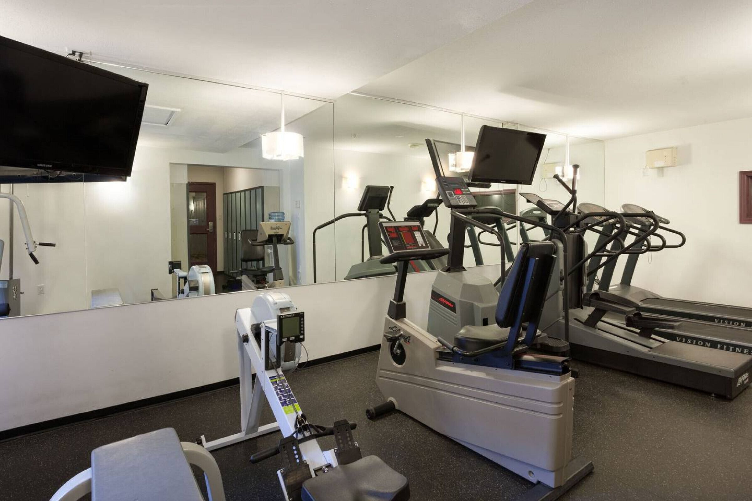 Ein Fitnessraum mit Laufband, Crosstrainer und Hantelrad, einem Fernseher und einem Spiegel.