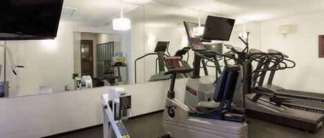 Ein Fitnessraum mit Laufband, Crosstrainer und Hantelrad, einem Fernseher und einem Spiegel.