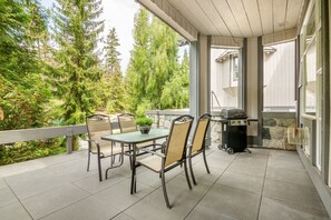 An outdoor patio with a table and chairs, a grill, and a view of trees. - Superb 2BR/2BA Steps to Gondola - AC + Gym! (Whistler)