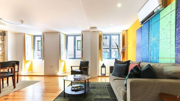 A spacious living room with a sofa, a coffee table, and a dining area with chairs. Large windows provide natural light and a view of the outside.