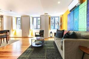 Ein geräumiges Wohnzimmer mit Sofa, Couchtisch und Essbereich mit Stühlen. Große Fenster sorgen für Tageslicht und bieten einen Ausblick ins Freie.