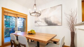 A dining area with a wooden table, four chairs, a chandelier, a large abstract painting, and a vase with branches. - Luxury 4BR - Private HT, Ski In/Out, Free Parking (Whistler)