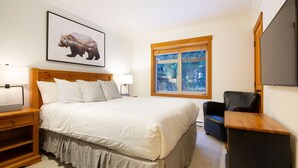 A bedroom with a large bed, a wooden headboard, a bedside table with a lamp, a chair, a window with a view of trees, and a bear picture on the wall. - Luxury 4BR - Private HT, Ski In/Out, Free Parking (Whistler)