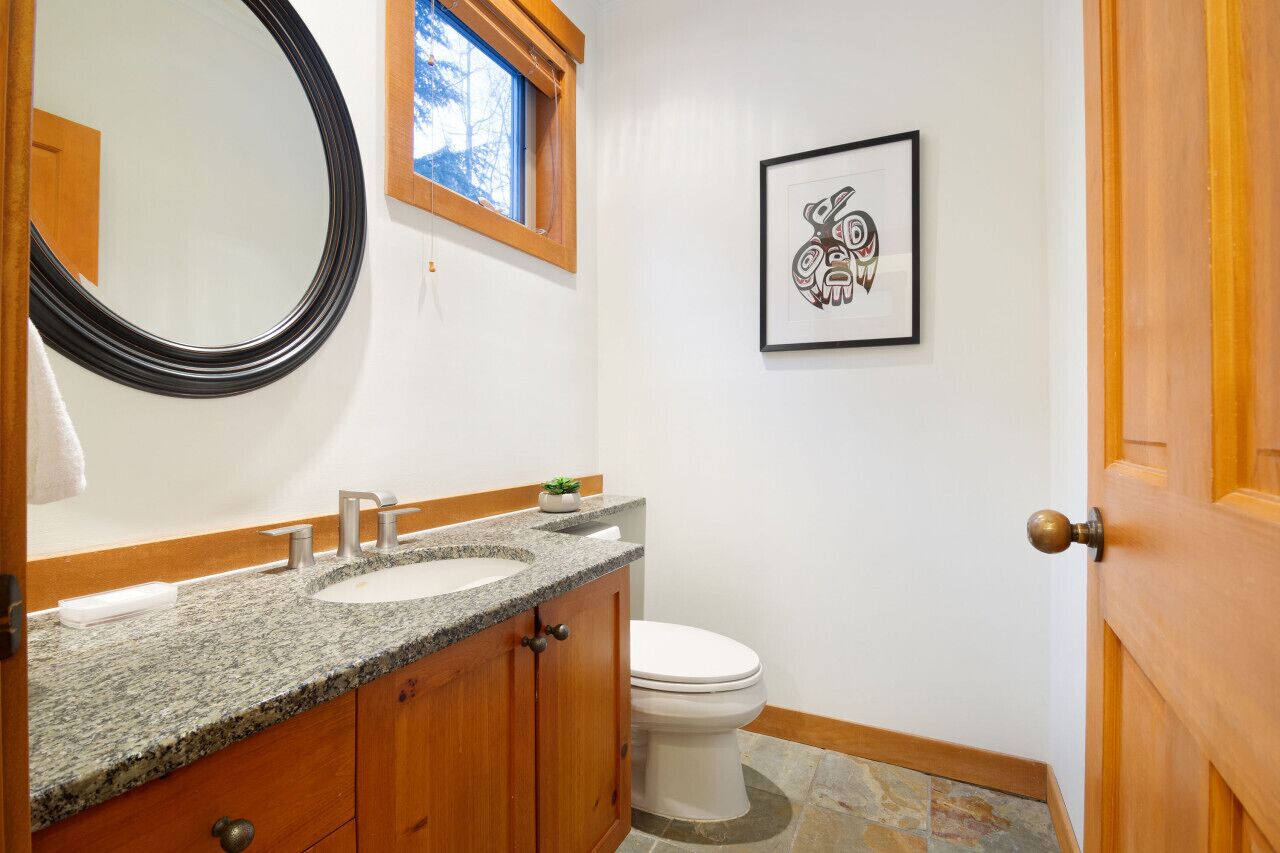 A bathroom with a granite countertop sink, wooden cabinets, a white toilet, a framed artwork on the wall, and a window above the sink.