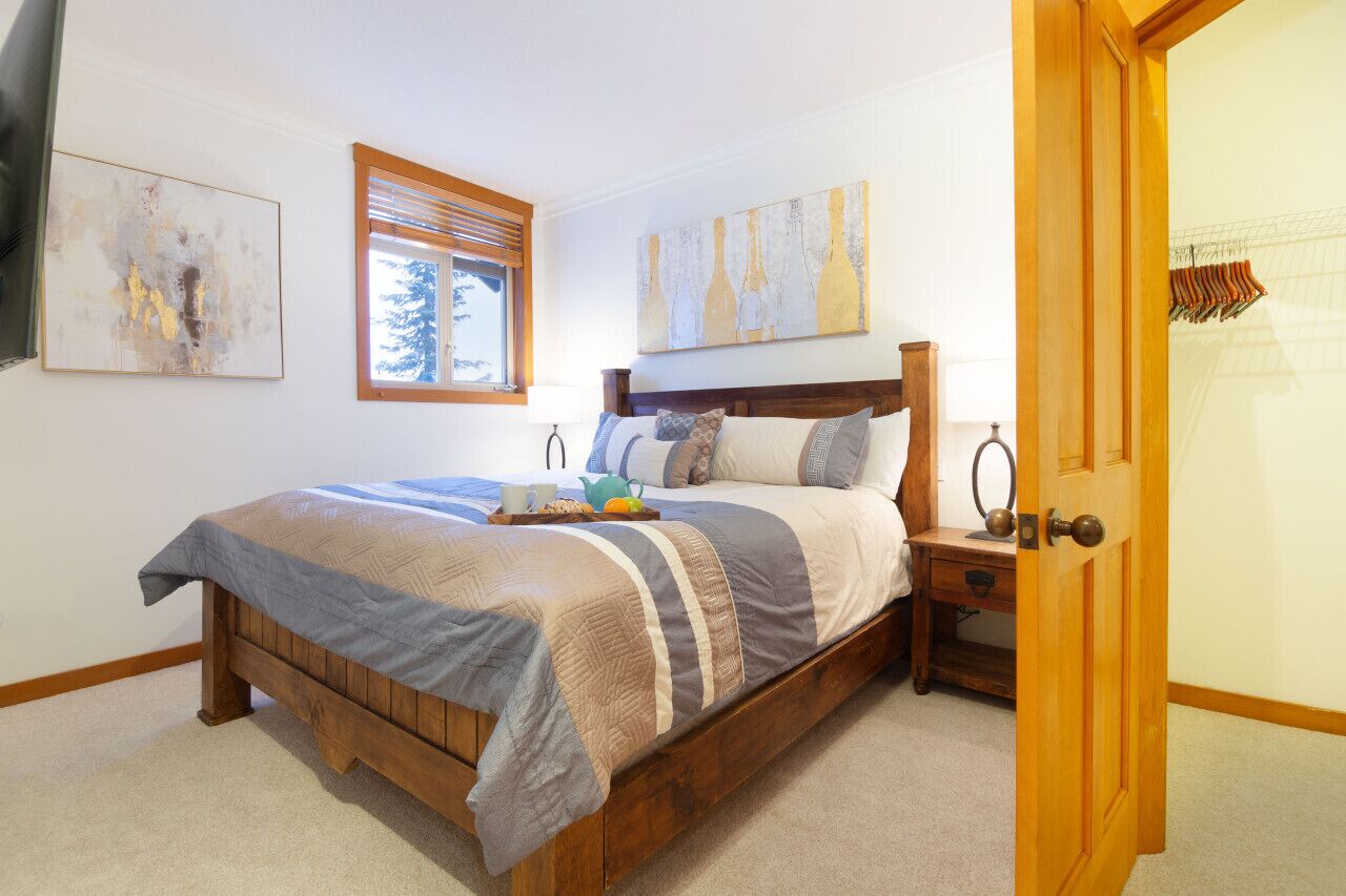 A bedroom with a wooden bed, a window with blinds, a bedside table with a lamp, and artwork on the walls.