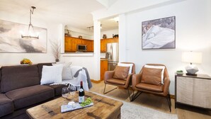 Un salon moderne avec un canapé marron, deux fauteuils en cuir, une table basse en bois sur laquelle se trouvent une bouteille de vin et des raisins, et un tableau représentant une montagne enneigée.
