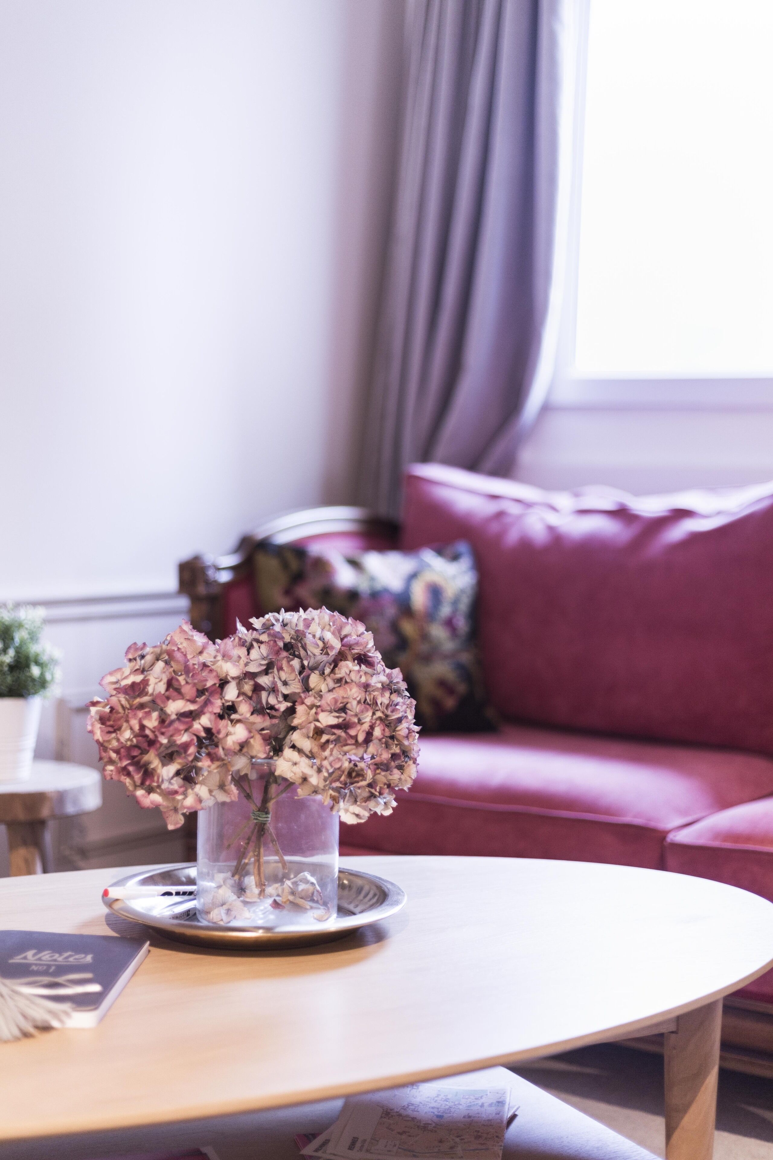 Una habitación con una mesa de centro redonda de madera, un jarrón con flores secas, un sofá rosa y una ventana con cortinas.