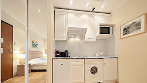 A compact hotel room with a kitchenette, a washing machine, a microwave, and a bed with a nightstand.