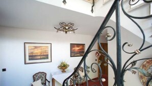 Un escalier orné de rampes en métal conduit à l’étage supérieur. Deux tableaux encadrés sont accrochés au mur, ainsi qu’une chaise et une table sur laquelle est posé un vase de fleurs.