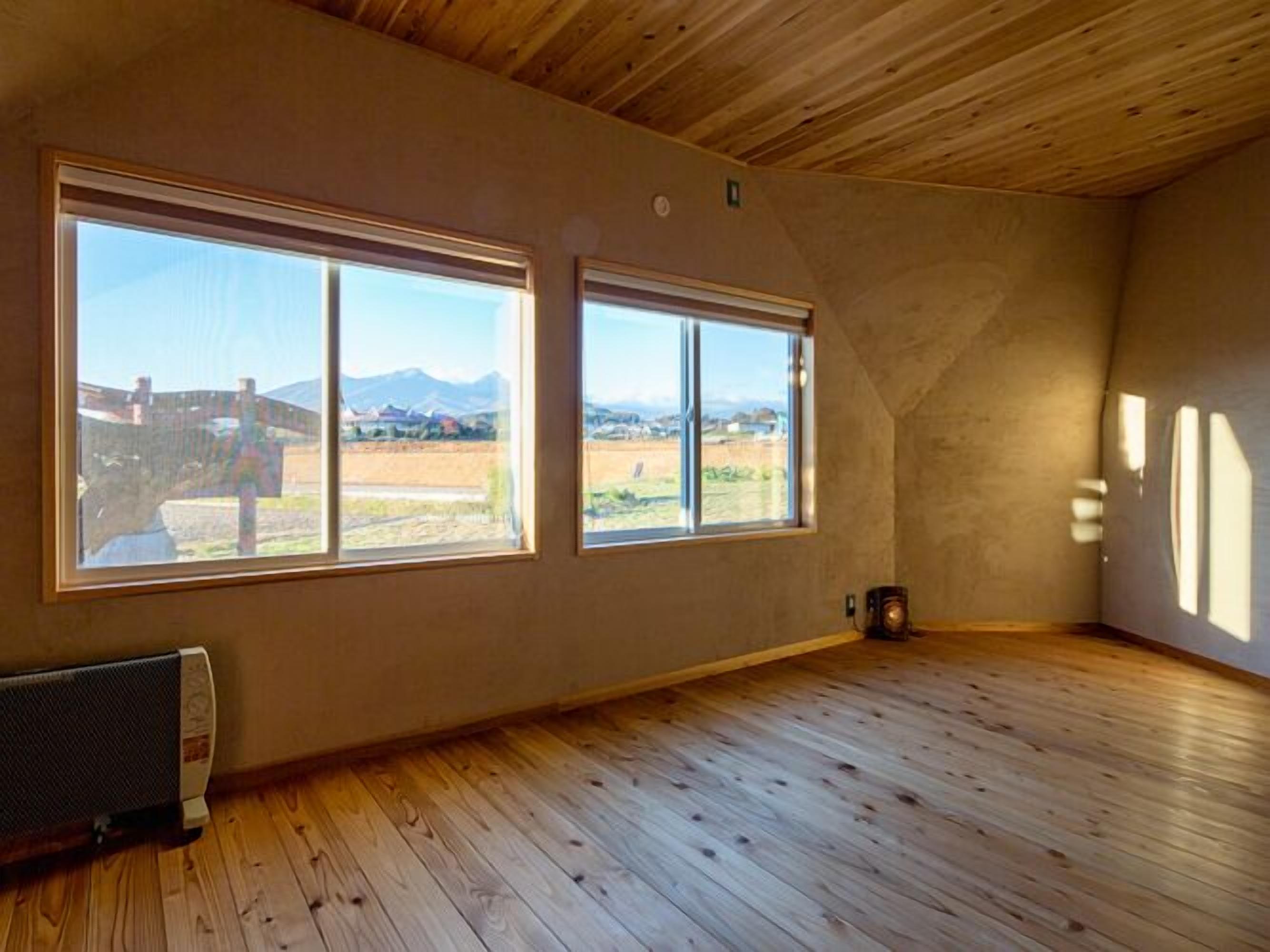 Ein Zimmer mit Holzdielen, zwei großen Fenstern mit Blick auf Berge und ein Feld sowie einer Heizung am Boden.