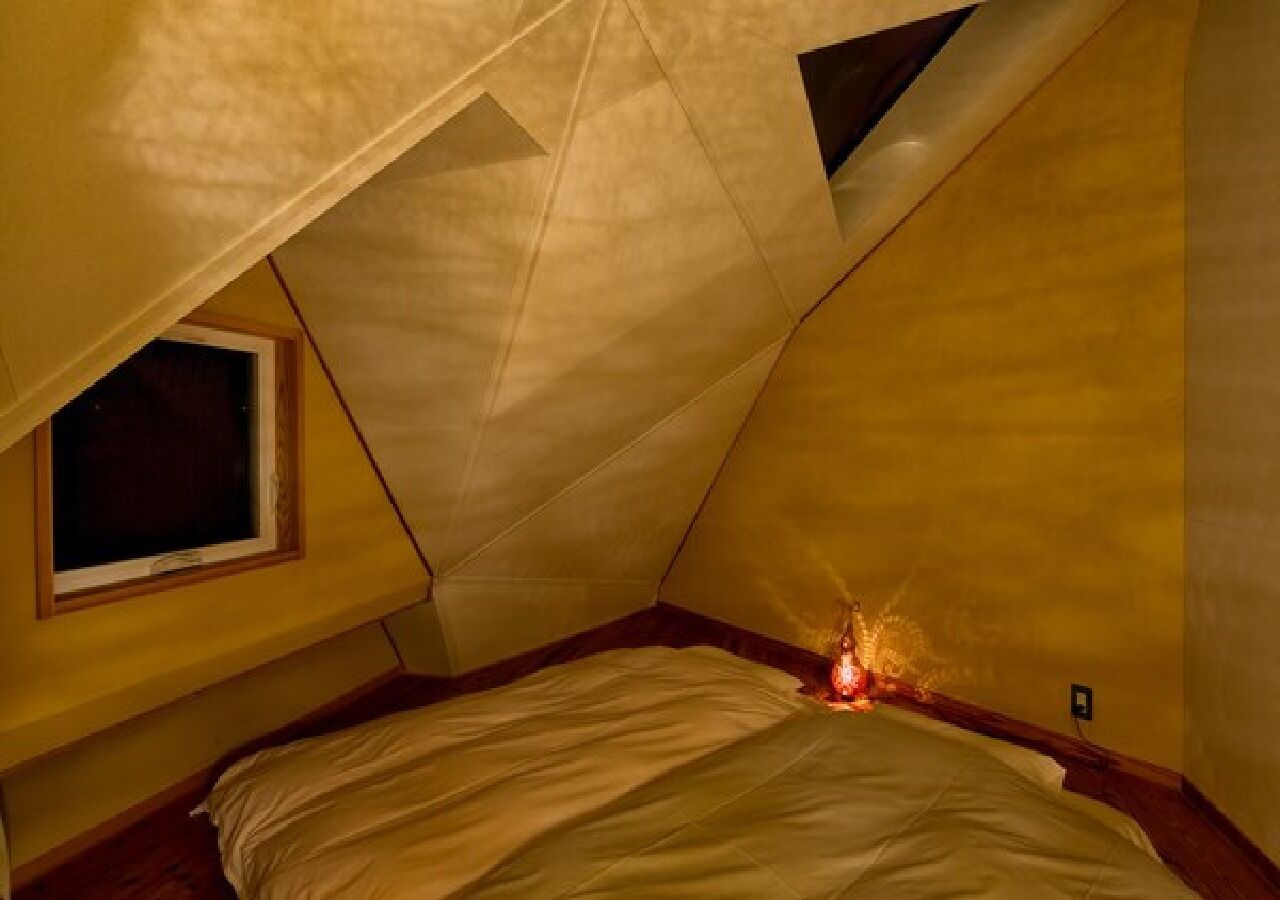 Ein quadratisches Schlafzimmer im Dachgeschoss mit einem einzelnen Fenster, einem Bett und einer sichtbaren Steckdose.