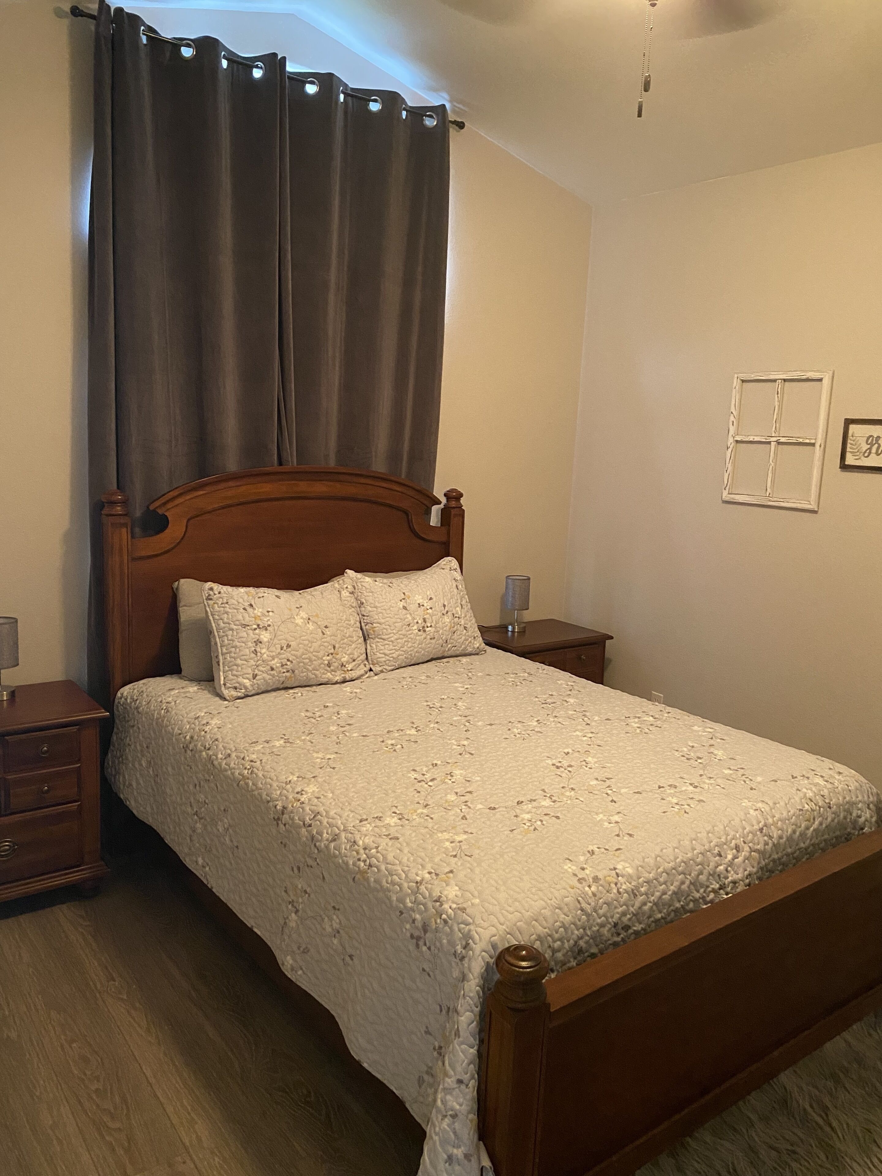 A bedroom with a wooden bed, a nightstand, a lamp, a window with a curtain, and a wall-mounted picture frame.