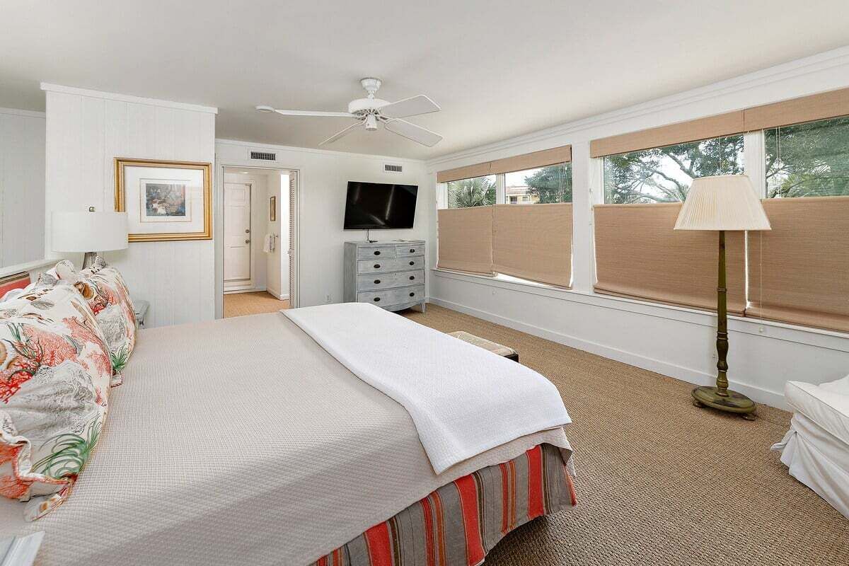 Ein Schlafzimmer mit Bett, Fernseher, Deckenventilator, Kommode und einem Fenster mit Jalousien.