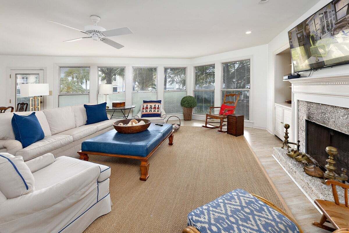 Ein geräumiges Wohnzimmer mit einem weißen Sofa, blauen Kissen, einem blauen Hocker, einem Kamin und einem darüber montierten Fernseher.