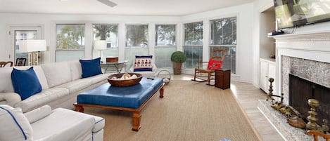 A spacious living room with a white sofa, blue cushions, a blue ottoman, a fireplace, and a television mounted above it.