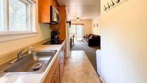 A kitchen with a double sink, microwave, and coffee maker. There is a dining area with a couch and a ceiling fan.