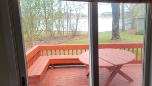 A wooden deck with a bench and a table, overlooking a grassy area with trees and a body of water.