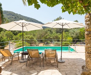 Zona de piscina con tres sombrillas, un pilar de piedra y vistas a un paisaje montañoso.