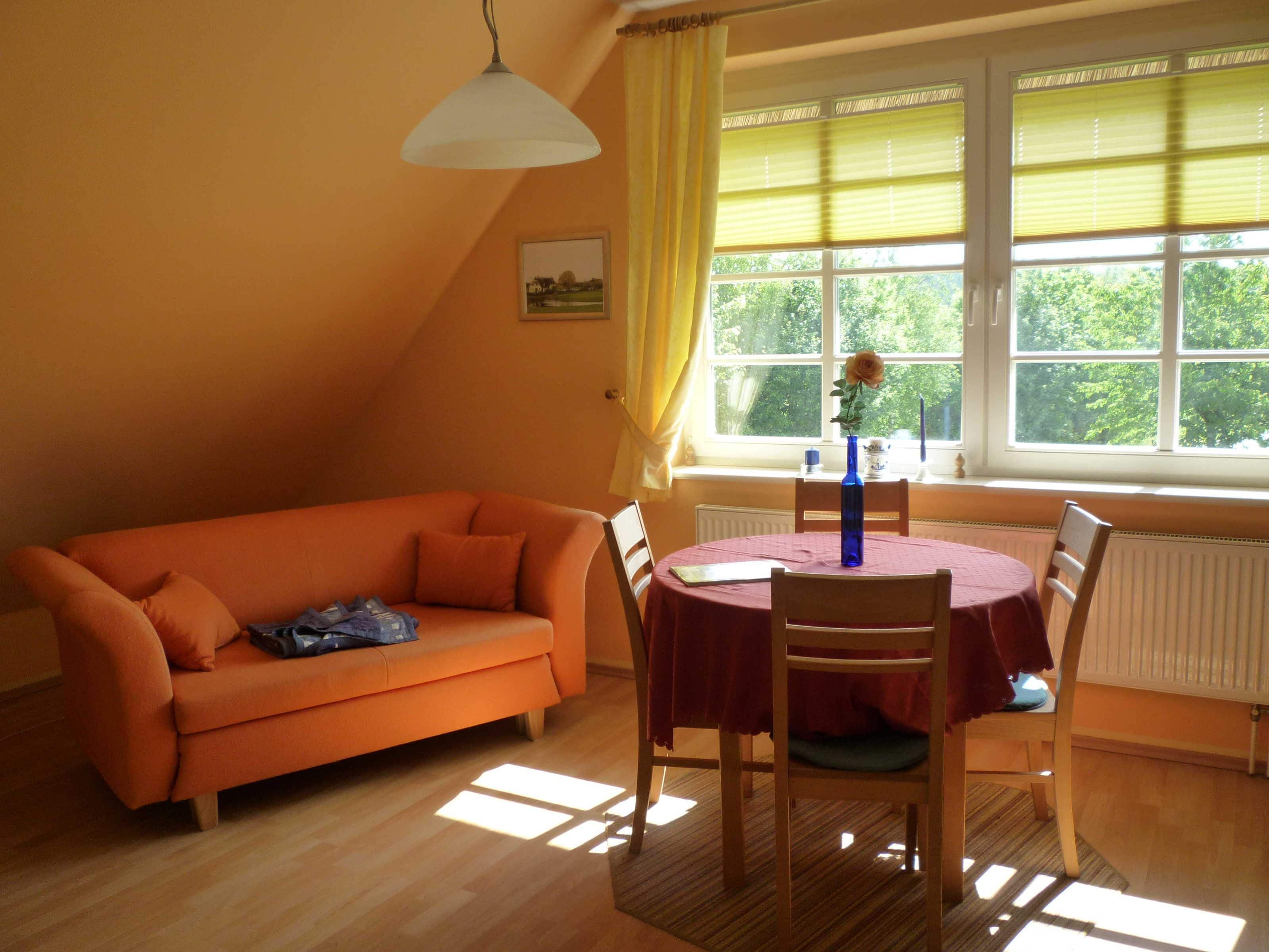 A room with an orange sofa, a dining table with chairs, a window with blinds, and a hanging light fixture.