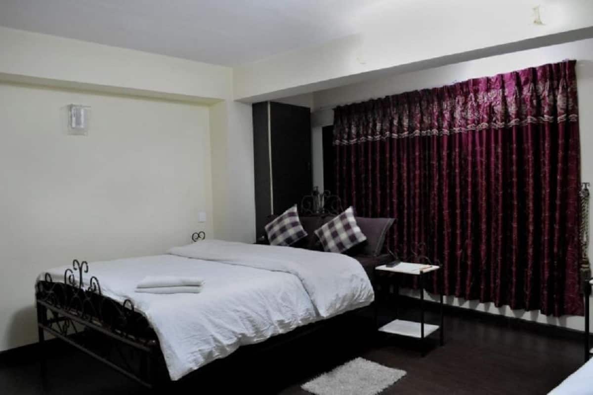 A bedroom with a bed, a nightstand, a chair, a window with curtains, and a wall-mounted light fixture.