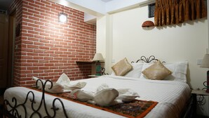 A hotel room with a brick wall, a bed with white bedding and decorative pillows, a nightstand with a lamp, and a window with curtains. - Cosy Hotel (Bhaktapur)