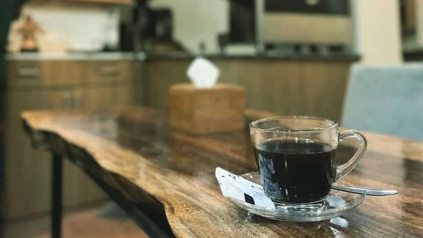 A cup of coffee on a wooden table in a kitchen with a coffee machine and shelves in the background. - Cosy Hotel (Bhaktapur)