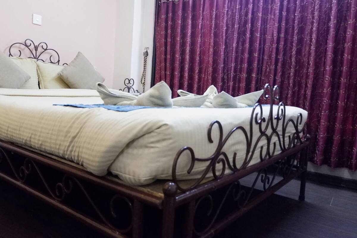 A neatly made bed with a white duvet and multiple pillows, a dark metal bed frame, and a purple curtain in the background.