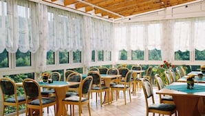 Un espacio exterior cubierto para comer con mesas y sillas de madera, ventanas amplias y vistas a zonas verdes.