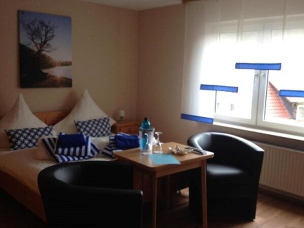 A room with a bed, two chairs, a table with a bottle and glasses, and a window with blinds.