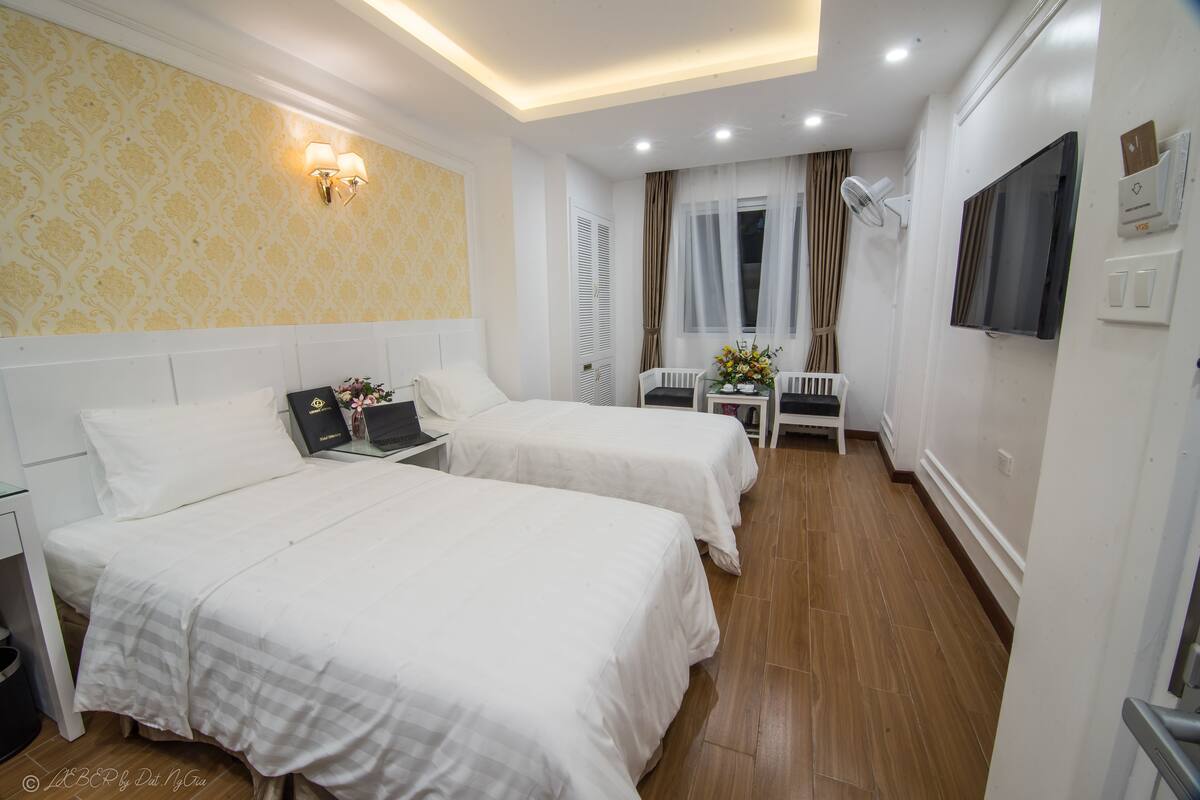 A hotel room with two beds, a desk with a laptop, a TV, and a small table with flowers.