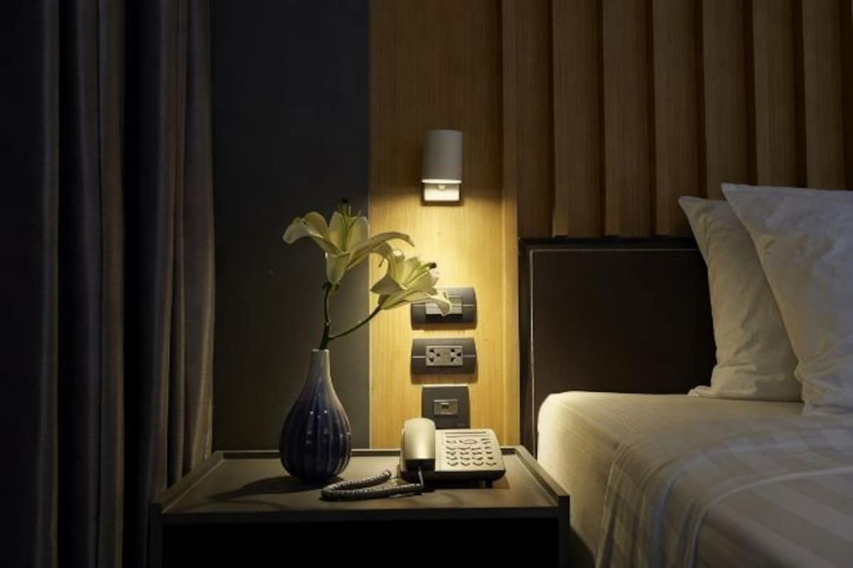 A hotel room with a bedside table, a vase with flowers, a bedside lamp, a phone, and a wall-mounted light.