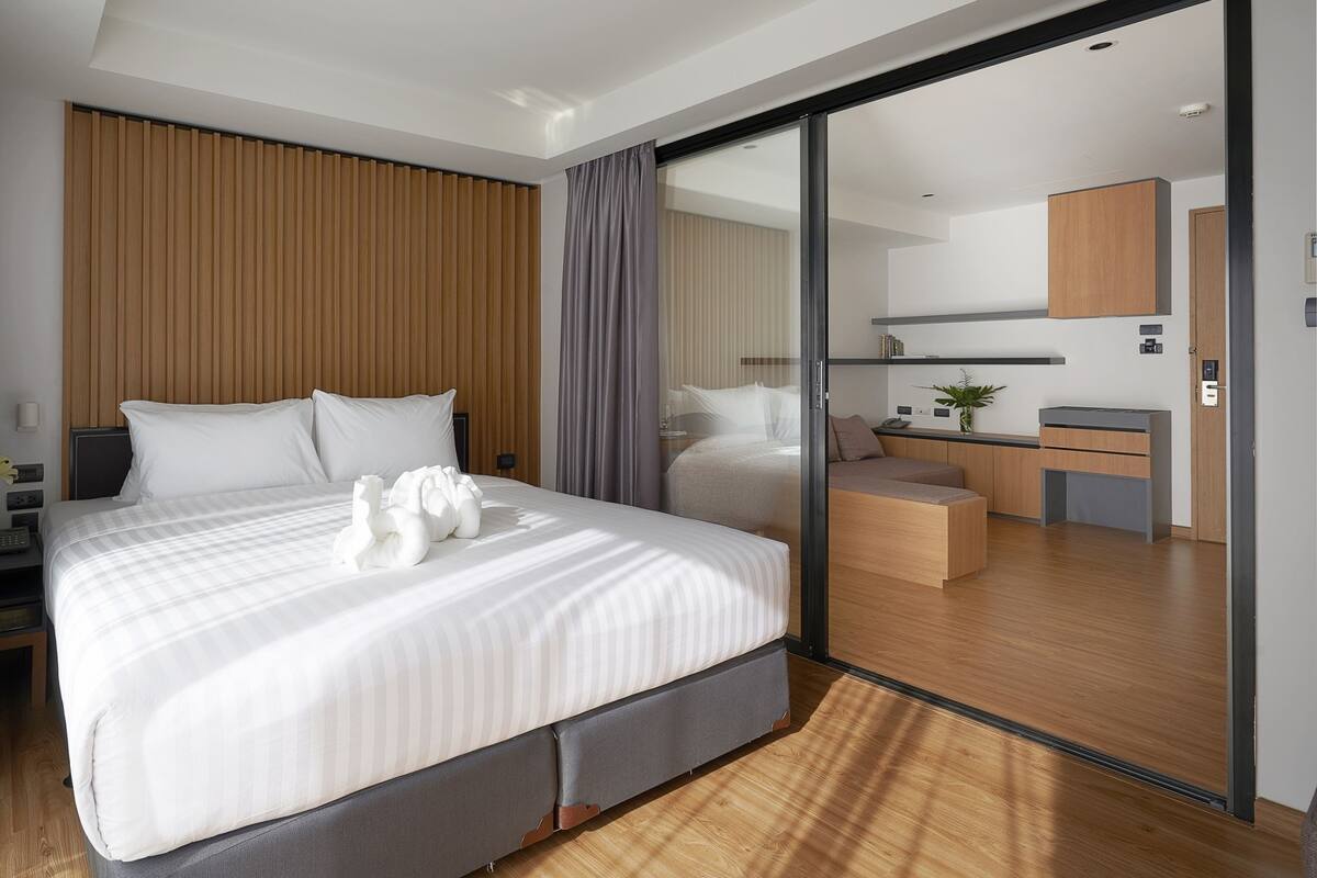 Une chambre d’hôtel moderne dotée d’un grand lit, d’une tête de lit en bois, d’une porte-fenêtre coulissante donnant sur un coin cuisine et d’un petit bureau avec une chaise.