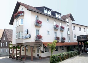 Un hotel de varios pisos con balcones decorados con flores, un cartel que indica que es una pensión y un estilo arquitectónico tradicional europeo.