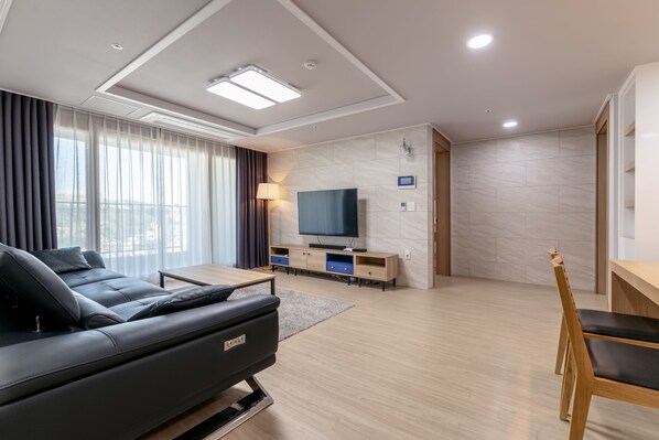 A modern living room with a sofa, TV, wooden furniture, and a city view. - Residence Hotel Eden Stay (Geoje)