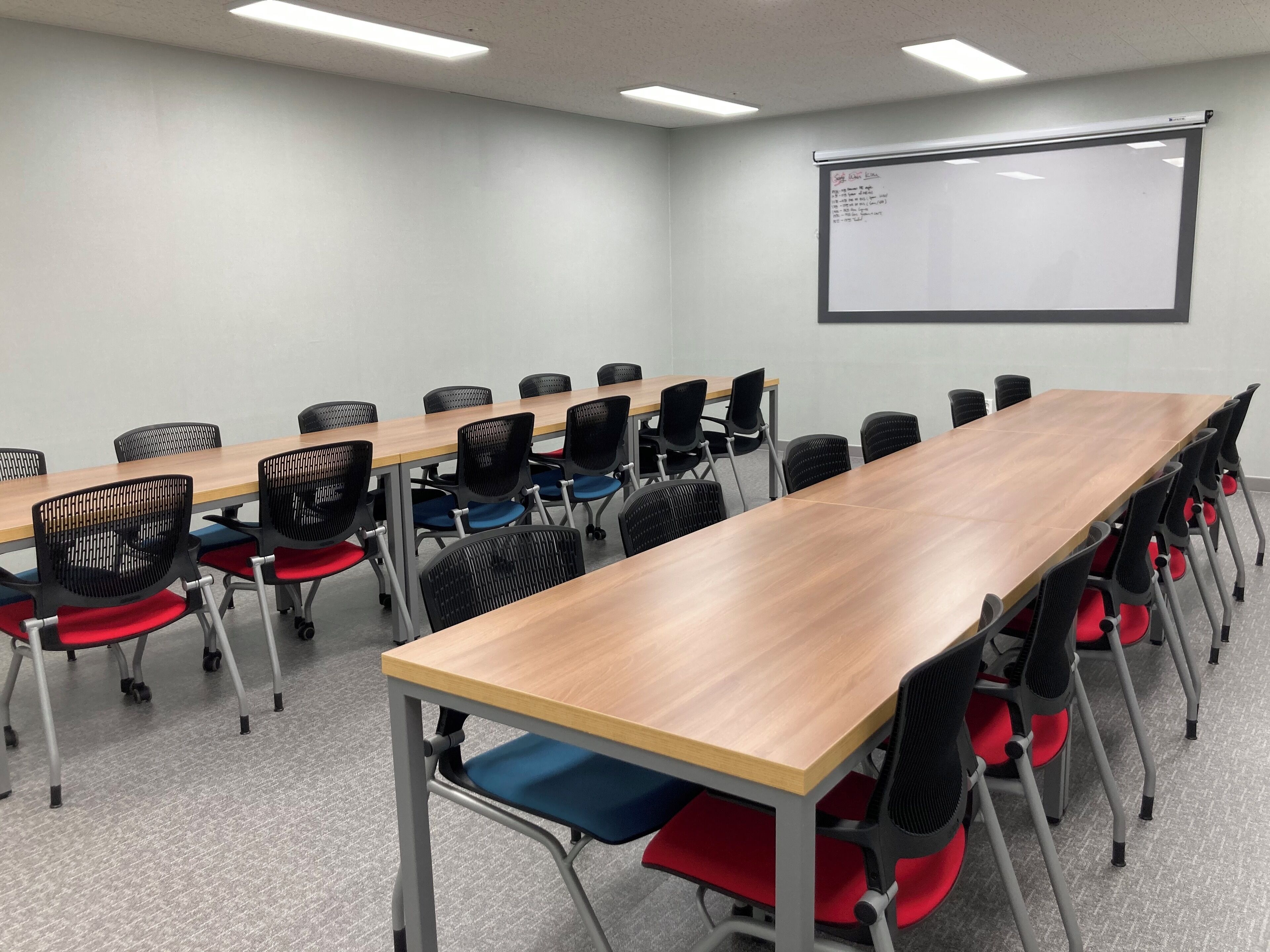 A conference room with a long table, multiple chairs, a whiteboard, and a projector screen.