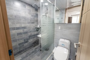 A modern bathroom with grey tiled walls, a glass-enclosed shower, a white toilet, and a wooden door. - Residence Hotel Eden Stay (Geoje)